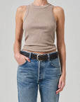 A person stands against a plain background, wearing the Isabel Rib Tank by Citizens Of Humanity/AGOLDE in beige, made from organic cotton, paired with blue jeans and a black woven belt. With one hand casually tucked in their pocket, they effortlessly showcase this versatile layering piece.