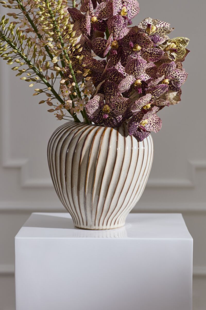 Tomar Vase from Accent Decor with purple orchids and greenery on a white pedestal against a pale wall.