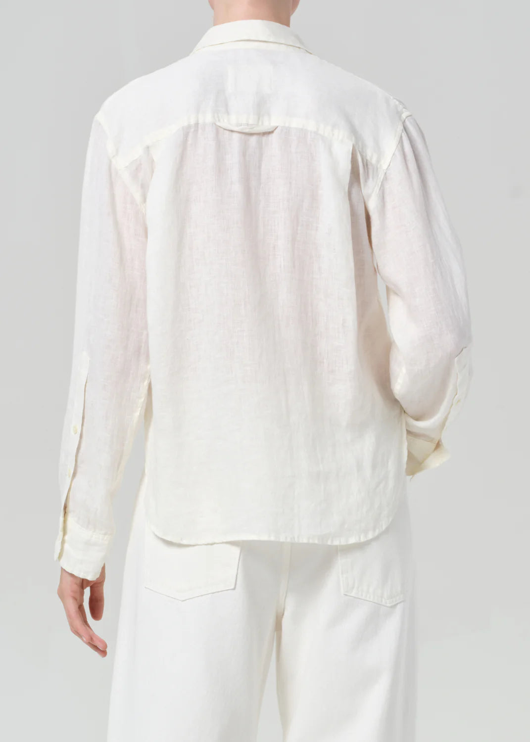 Someone stands facing away, showcasing the relaxed silhouette of the Citizens Of Humanity/AGOLDE kayla shirt in off-white Japanese shirting, paired with light pants against a plain background.