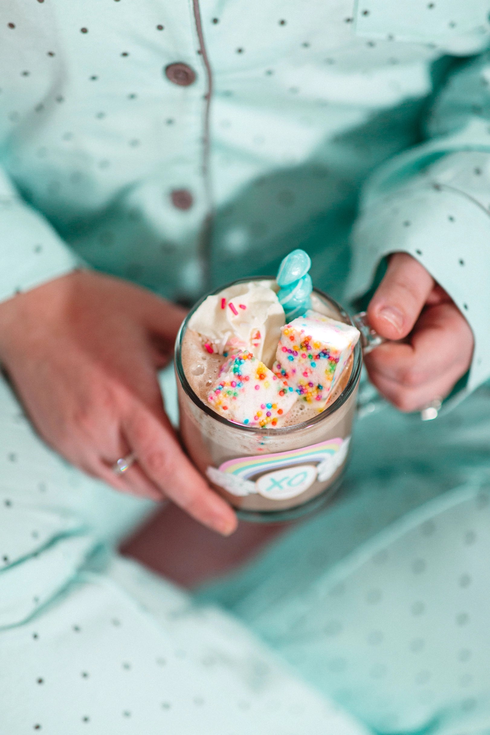 Wearing light blue polka-dot pajamas, a person holds a mug of hot chocolate with whipped cream, Faire Funfetti Marshmallows, and rainbow sprinkles. The mug’s bright XO design adds extra color to the scene.