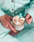 Wearing light blue polka-dot pajamas, a person holds a mug of hot chocolate with whipped cream, Faire Funfetti Marshmallows, and rainbow sprinkles. The mug’s bright XO design adds extra color to the scene.