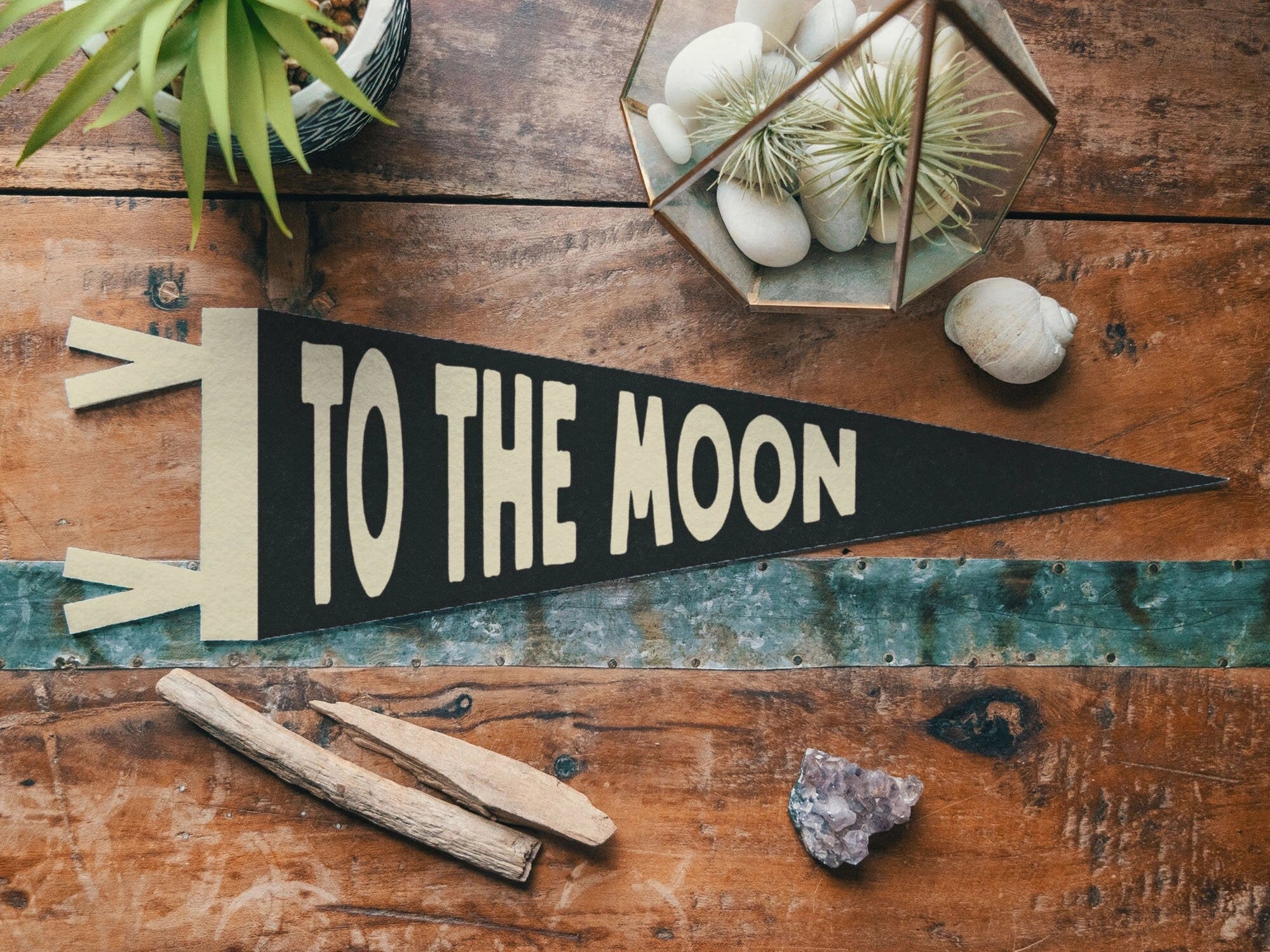 The Faire To The Moon Felt Pennant, featuring large cream letters, sits on a rustic table amid plants and crystals—ideal as vintage nursery decor or a charming family love banner.