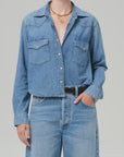 A person poses western style with one hand in a pocket, wearing a Citizens Of Humanity/AGOLDE cropped western shirt in light blue denim with two chest pockets and matching high-waisted jeans, accessorized with layered gold necklaces and a black belt.