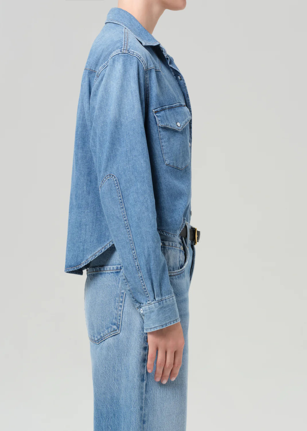 A person wearing the Citizens Of Humanity/AGOLDE cropped western shirt with flap chest pockets and a matching skirt is shown in profile against a light gray background, with their head partially out of frame.