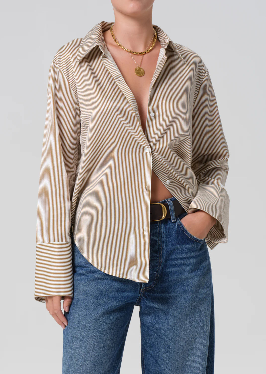 A person wearing the beige and white striped Camilia Shirt by Citizens Of Humanity/AGOLDE, partly unbuttoned, styled with blue jeans, a black belt, and layered gold necklaces, stands against a plain light background.