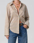 A person wearing the beige and white striped Camilia Shirt by Citizens Of Humanity/AGOLDE, partly unbuttoned, styled with blue jeans, a black belt, and layered gold necklaces, stands against a plain light background.