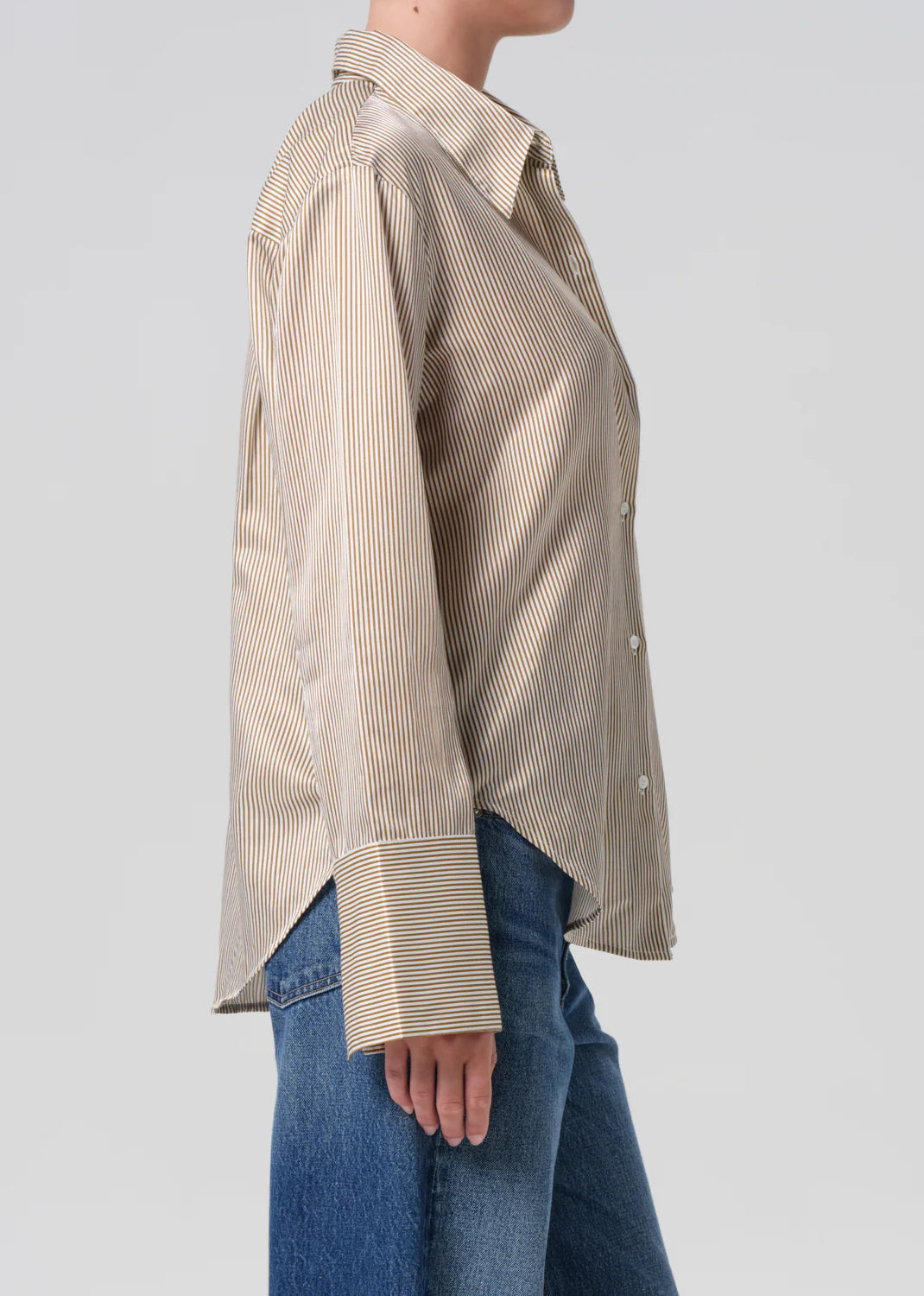 A person in the Citizens Of Humanity/AGOLDE Camilia Shirt—beige and white vertical stripes, long sleeves with wide cuffs—and blue jeans is shown from the side against a plain light background.