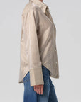 A person in the Citizens Of Humanity/AGOLDE Camilia Shirt—beige and white vertical stripes, long sleeves with wide cuffs—and blue jeans is shown from the side against a plain light background.