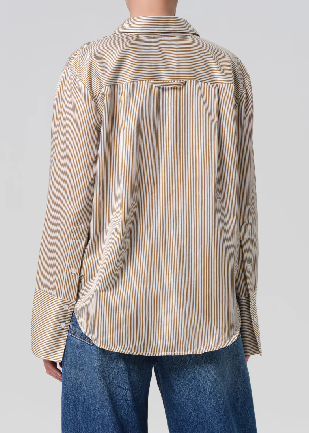 A person is wearing the Citizens Of Humanity/AGOLDE Camilia Shirt—a loose-fitting, beige and white striped cotton silk blend blouse with buttoned cuffs—paired with blue jeans. Photo shows the back view against a plain background.