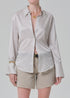 Wearing the camilia shirt S26 by Citizens Of Humanity/AGOLDE—a shiny beige striped cotton-silk button-down with flared cuffs—paired with beige shorts, a black belt, gold necklaces, and a bracelet against a light gray background.