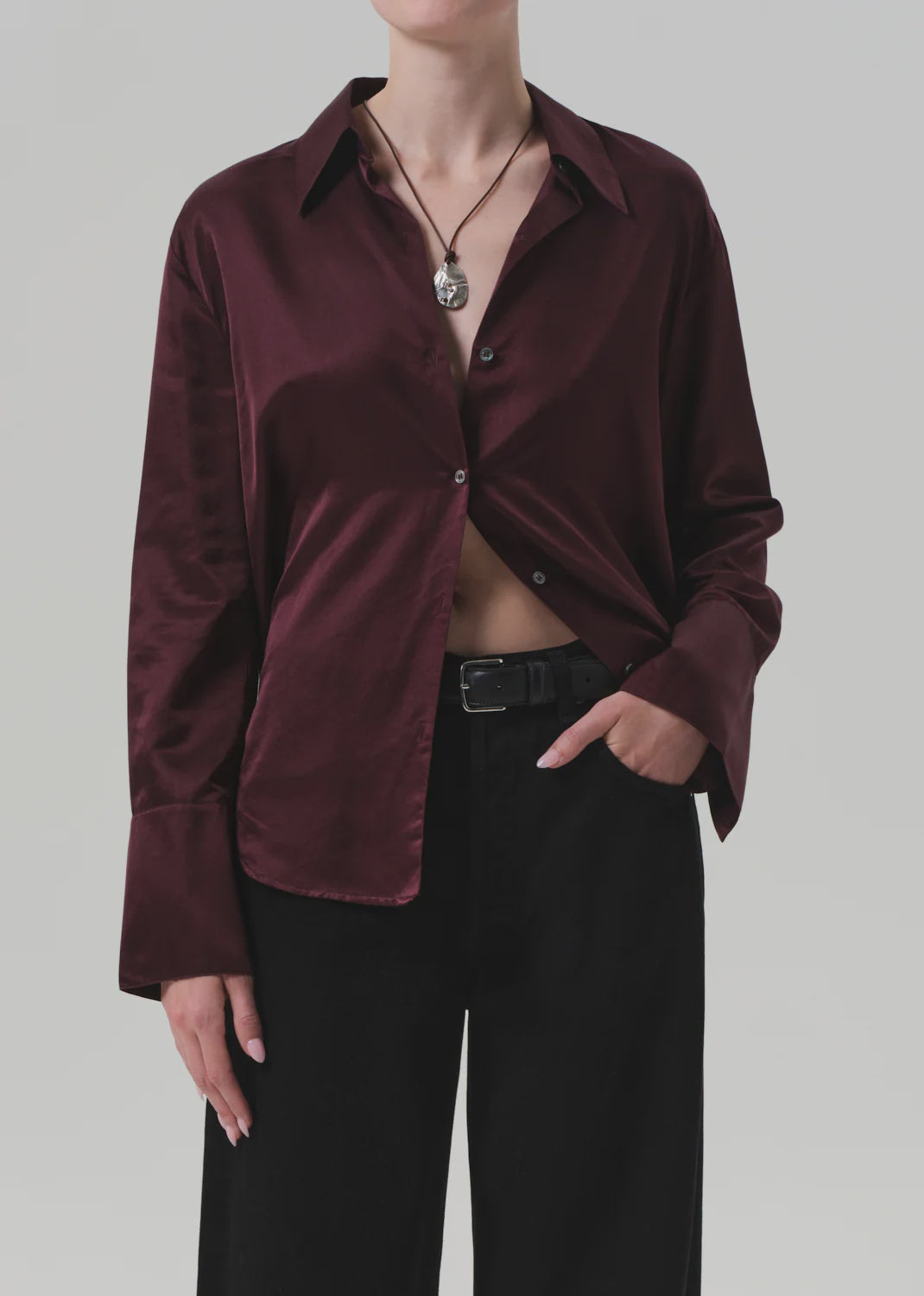 A model wears the Camilia Shirt by Citizens Of Humanity/AGOLDE in shiny burgundy cotton-silk blend, partially unbuttoned, with black wide-leg pants, a black belt, and a pendant necklace against a plain light gray background.