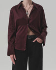 A model wears the Camilia Shirt by Citizens Of Humanity/AGOLDE in shiny burgundy cotton-silk blend, partially unbuttoned, with black wide-leg pants, a black belt, and a pendant necklace against a plain light gray background.
