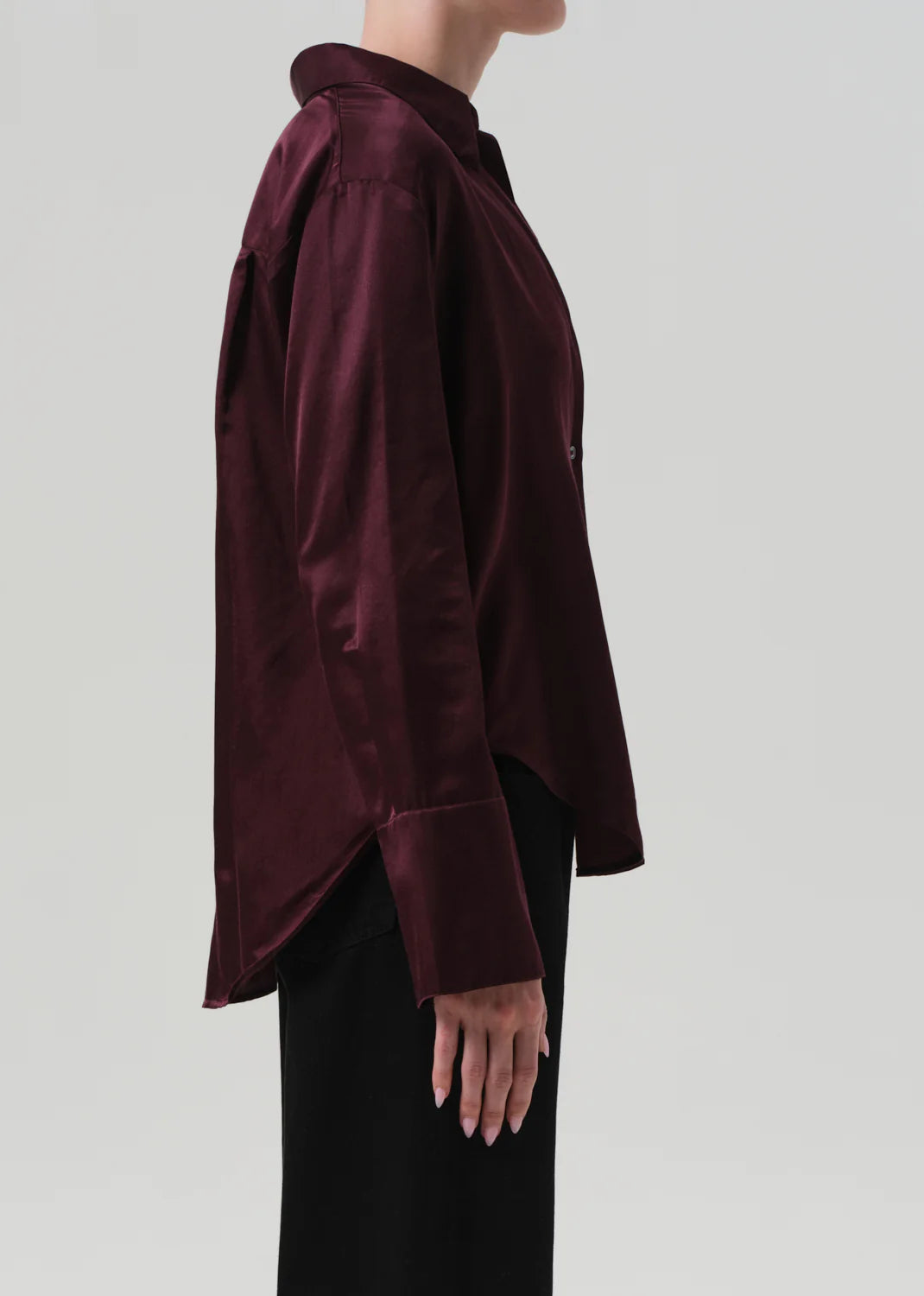 A person stands in profile against a plain background, wearing the Citizens Of Humanity/AGOLDE Camilia Shirt—a long sleeve, dark burgundy, cotton silk blend button-front blouse with a collar and a slightly loose fit that extends past the hips—paired with black pants.