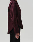 A person stands in profile against a plain background, wearing the Citizens Of Humanity/AGOLDE Camilia Shirt—a long sleeve, dark burgundy, cotton silk blend button-front blouse with a collar and a slightly loose fit that extends past the hips—paired with black pants.