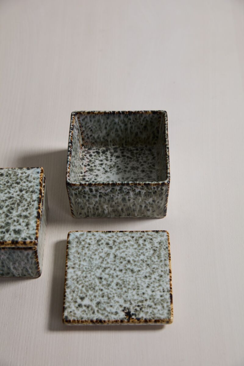 The Whisper Lidded Box by Accent Decor, featuring a square, speckled ceramic design in gray and brown, sits on a light surface with its matching lid beside it—ideal as stylish jewelry storage or a decorative home accent.