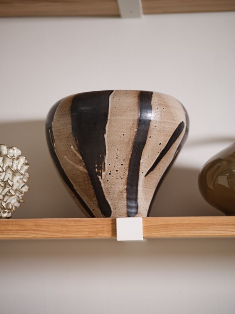 The Atwood Vase by Accent Decor—a hand painted ceramic piece with a beige base and bold vertical black brushstrokes—rests on a wooden shelf, surrounded by sculptural vessels and decorative objects.