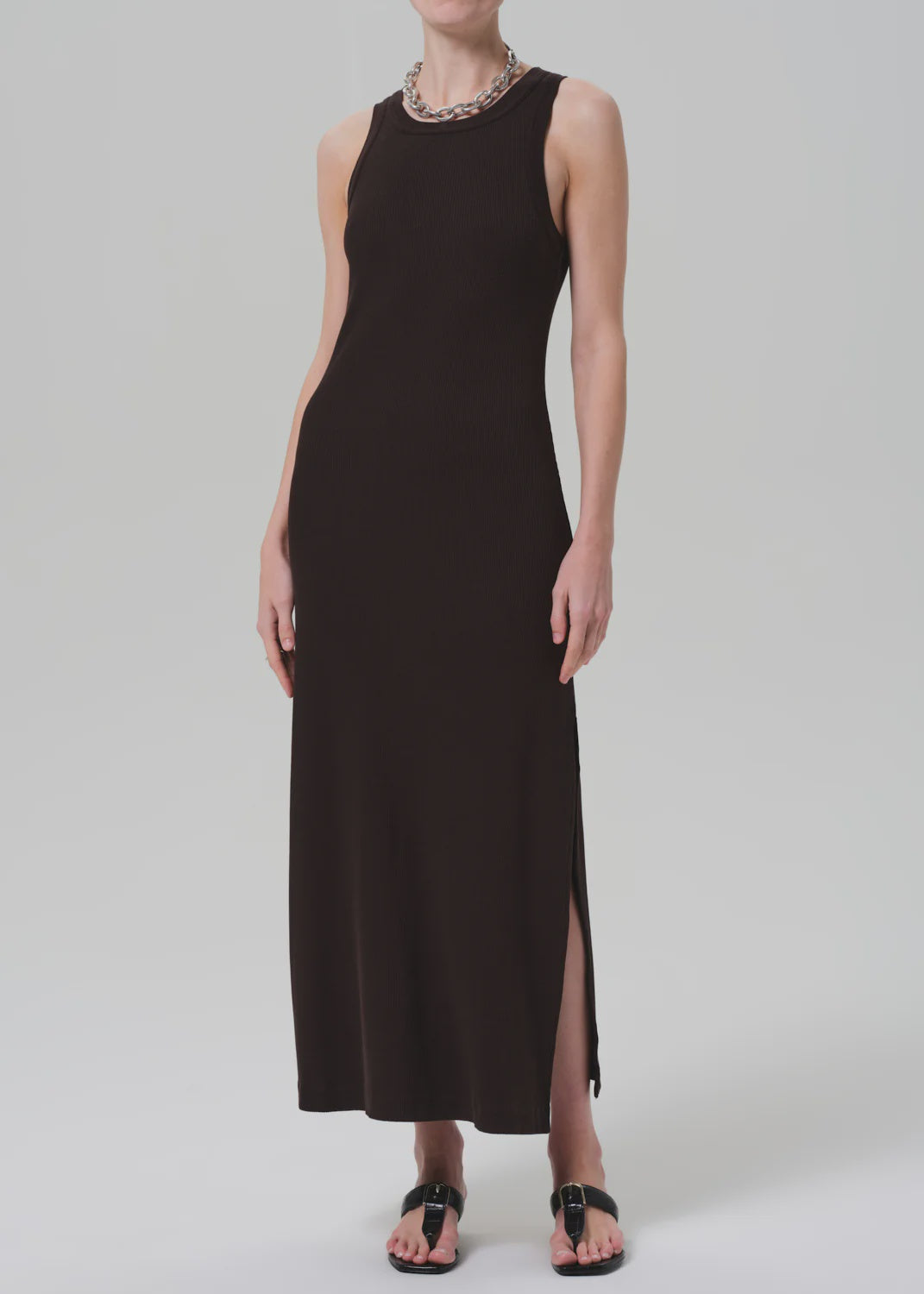 A person wears the Isabel Tank Dress by Citizens Of Humanity/AGOLDE—a sleeveless black midi with side slit—styled with a chunky chain necklace and black sandals, standing against a plain, light-colored background.