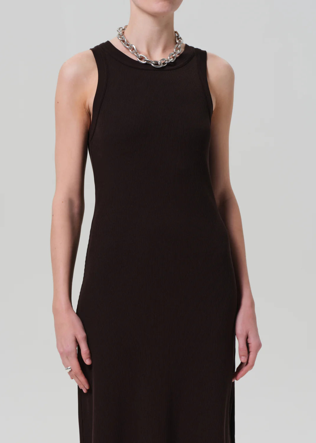 A person wears the Isabel Tank Dress by Citizens Of Humanity/AGOLDE—a sleeveless, fitted black ribbed dress—with a chunky silver chain necklace. Their hand rests by their side, and the plain light background highlights the cropped composition.