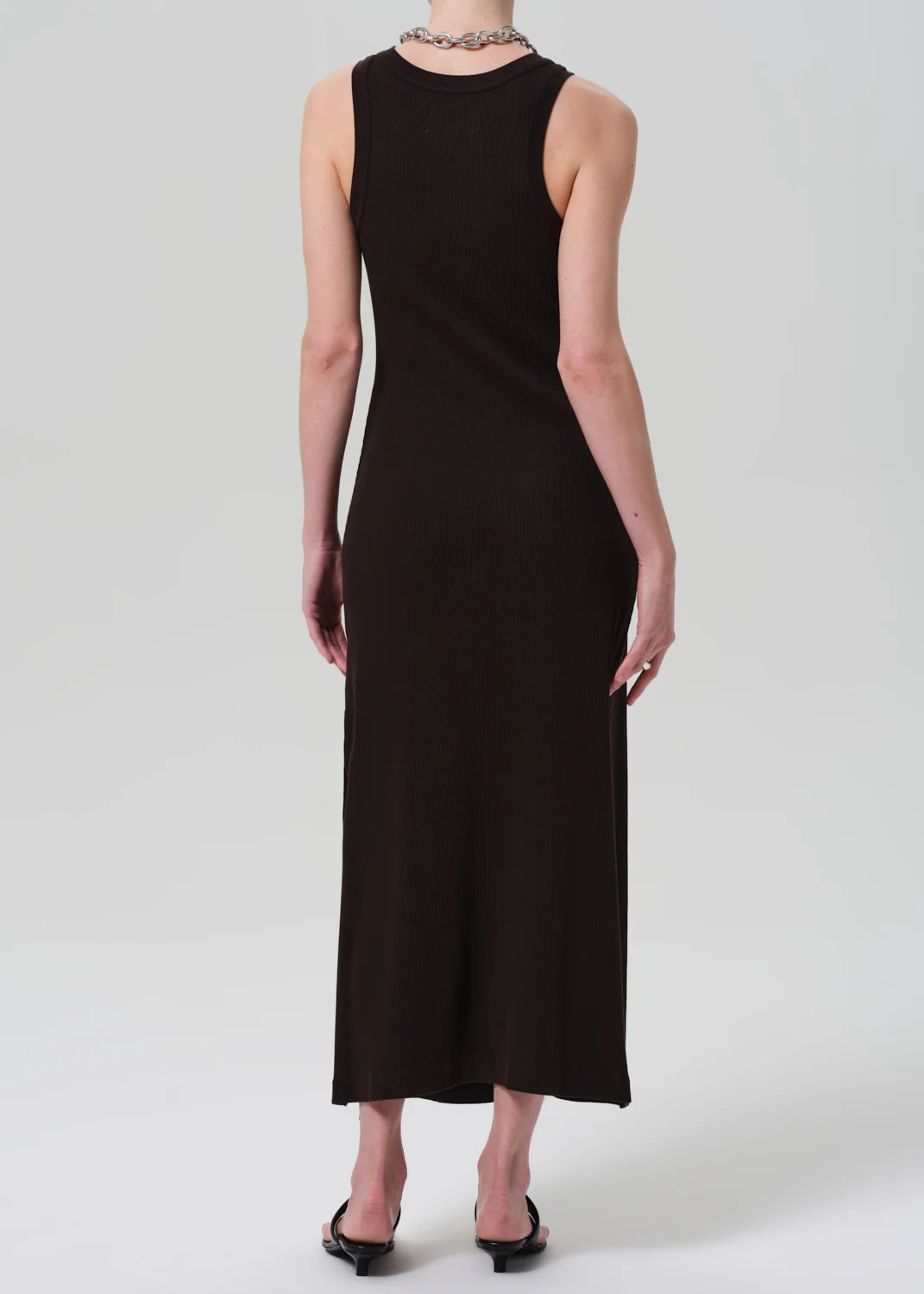 A person stands facing away, wearing the ankle-length black Isabel Tank Dress by Citizens Of Humanity/AGOLDE and black open-toe heels, accented with a chunky necklace against a plain, light background.