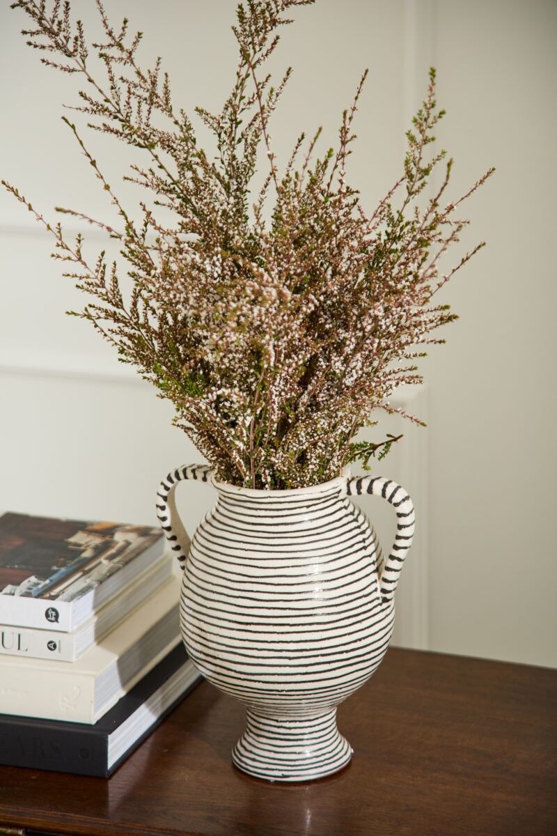 The Lines Vase by Accent Decor, a striped ceramic piece with two handles, displays long-stemmed greenery and white flowers. Handcrafted, it rests on a wooden surface beside stacked books.