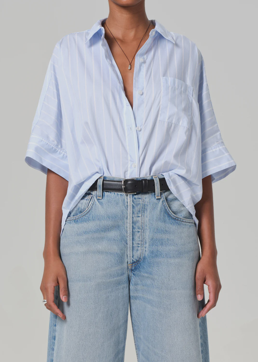 The person wears a Claire Origami Stripe shirt from Citizens Of Humanity/AGOLDE, featuring light blue and white stripes. It's tucked into high-waisted jeans with a black belt, paired with a necklace and ring. This stylish outfit, made of regenerative cotton, stands out against the gray background.