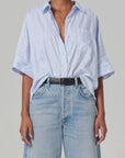 The person wears a Claire Origami Stripe shirt from Citizens Of Humanity/AGOLDE, featuring light blue and white stripes. It's tucked into high-waisted jeans with a black belt, paired with a necklace and ring. This stylish outfit, made of regenerative cotton, stands out against the gray background.
