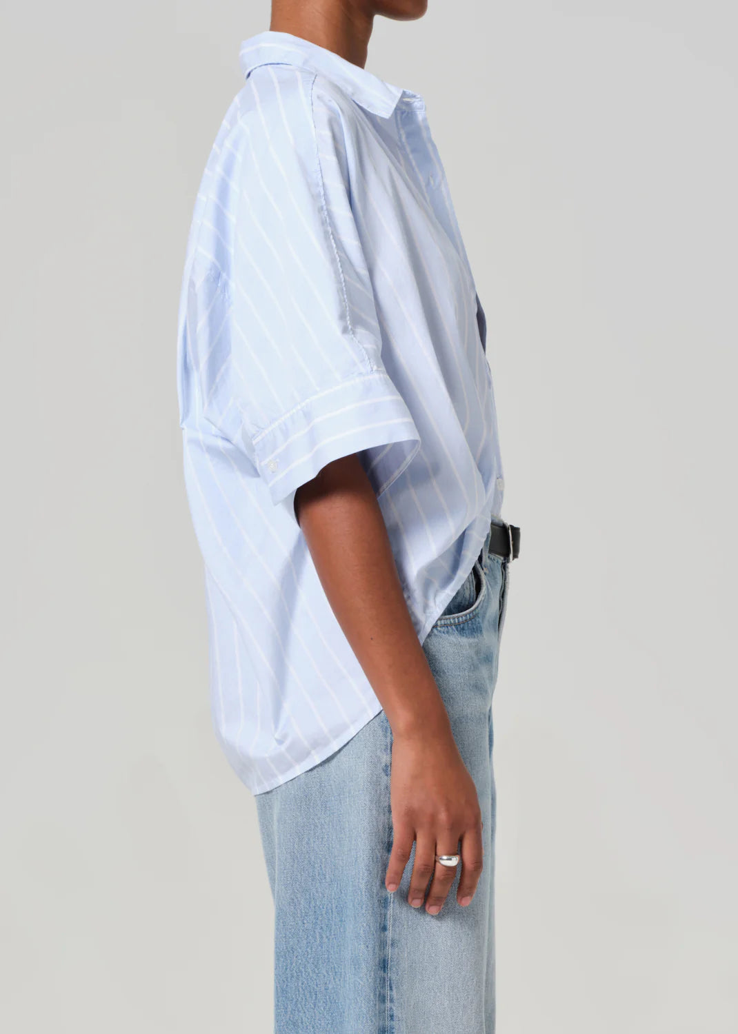 Wearing the Citizens Of Humanity/AGOLDE Claire Origami Stripe, a loose-fit light blue and white striped button-up paired with light blue jeans, this person exudes a relaxed style. Their hand, adorned with a ring, rests beside them against a plain gray backdrop. The ensemble is crafted from regenerative cotton.