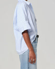 Wearing the Citizens Of Humanity/AGOLDE Claire Origami Stripe, a loose-fit light blue and white striped button-up paired with light blue jeans, this person exudes a relaxed style. Their hand, adorned with a ring, rests beside them against a plain gray backdrop. The ensemble is crafted from regenerative cotton.