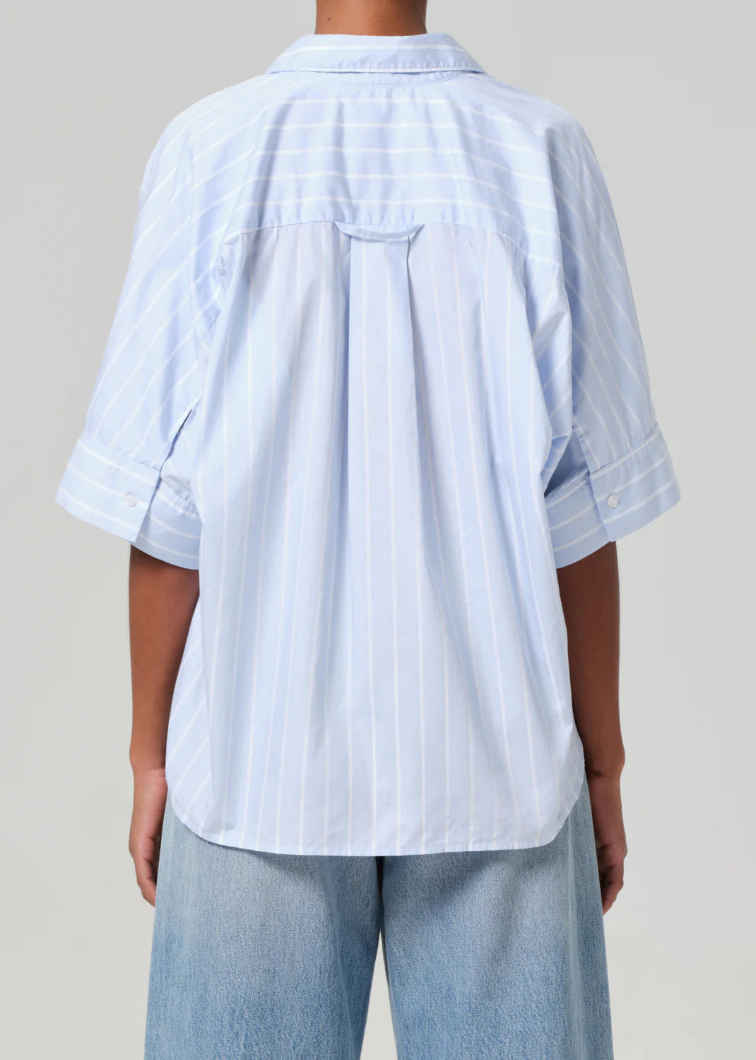 Rear view showing a person in a light blue, Claire Origami Stripe shirt from Citizens Of Humanity/AGOLDE, short-sleeved and loose fit, paired with light blue jeans. The regenerative cotton fabric drapes gently over the shoulders against a plain gray background.