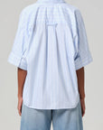 Rear view showing a person in a light blue, Claire Origami Stripe shirt from Citizens Of Humanity/AGOLDE, short-sleeved and loose fit, paired with light blue jeans. The regenerative cotton fabric drapes gently over the shoulders against a plain gray background.