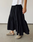 A person wearing a white t-shirt, Hey Gang’s The Ranch Skirt in black with ruffles, and beige slip-on clogs stands on a gray floor against a plain light-colored wall.