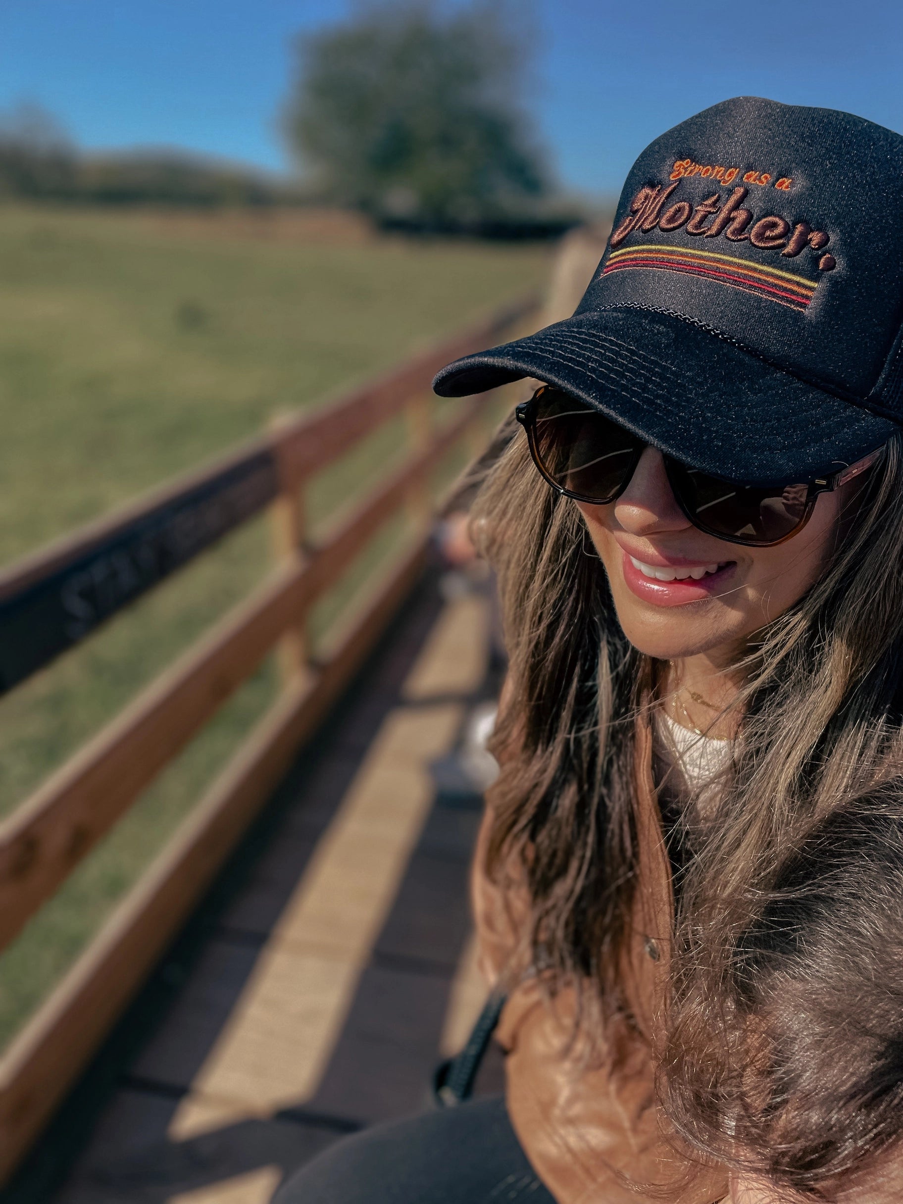 A woman in sunglasses smiles while walking on a sunny wooden bridge, wearing the Faire Retro Mother Hat with "Strong as a Mother" on the front. Green grass and trees are visible in the background.