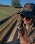 A woman in sunglasses smiles while walking on a sunny wooden bridge, wearing the Faire Retro Mother Hat with "Strong as a Mother" on the front. Green grass and trees are visible in the background.