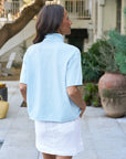 A woman with long dark hair and gray streaks stands outdoors in a patio or garden, wearing the Frank & Eileen Patty Popover SS Henley in light blue and a white skirt, her hands tucked into her pockets as she faces away.