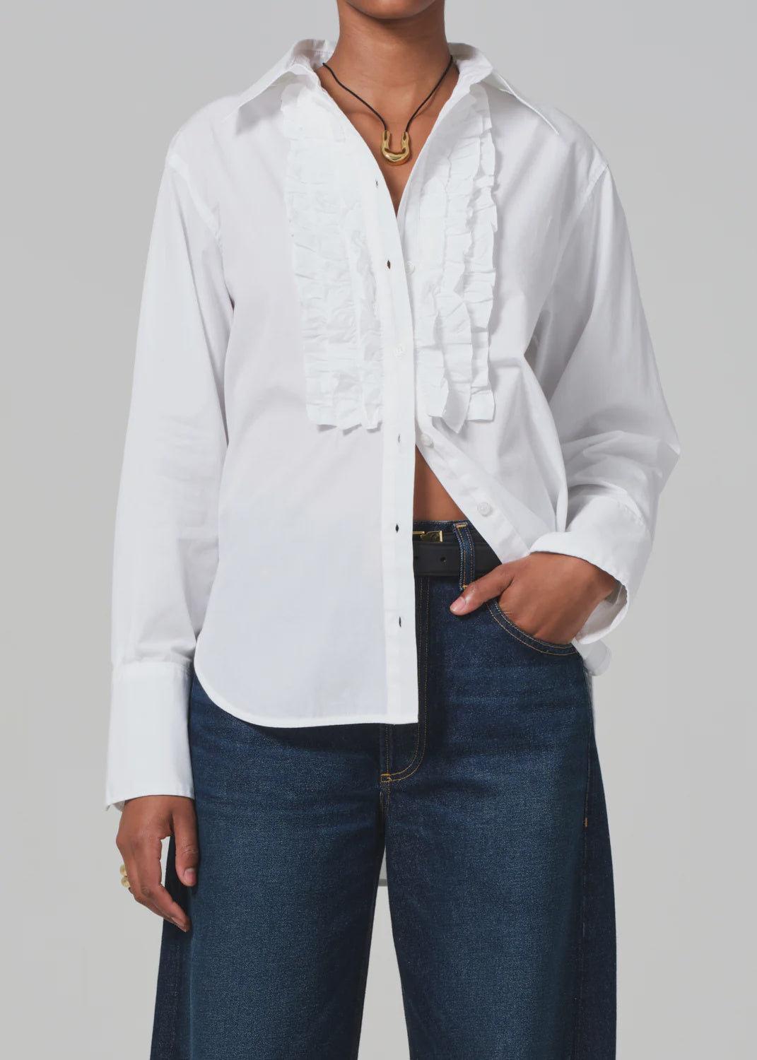 A person wearing the Ruffle Tuxedo Shirt by Citizens Of Humanity/AGOLDE, paired with dark blue jeans. Their left hand is in the pocket, and they are accessorized with a necklace featuring a pendant. The background is plain light gray.