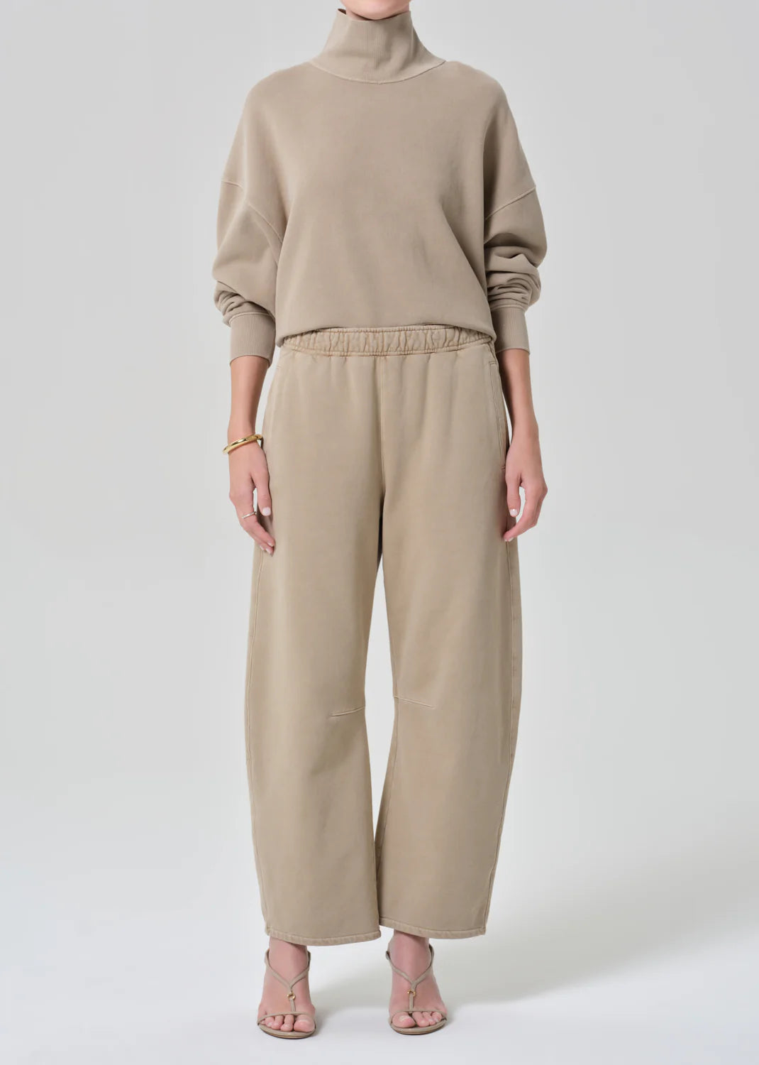 A person wearing Citizens Of Humanity/AGOLDE's miro sweatpant with a beige turtleneck, light sandals, and a gold bracelet stands against a plain background.