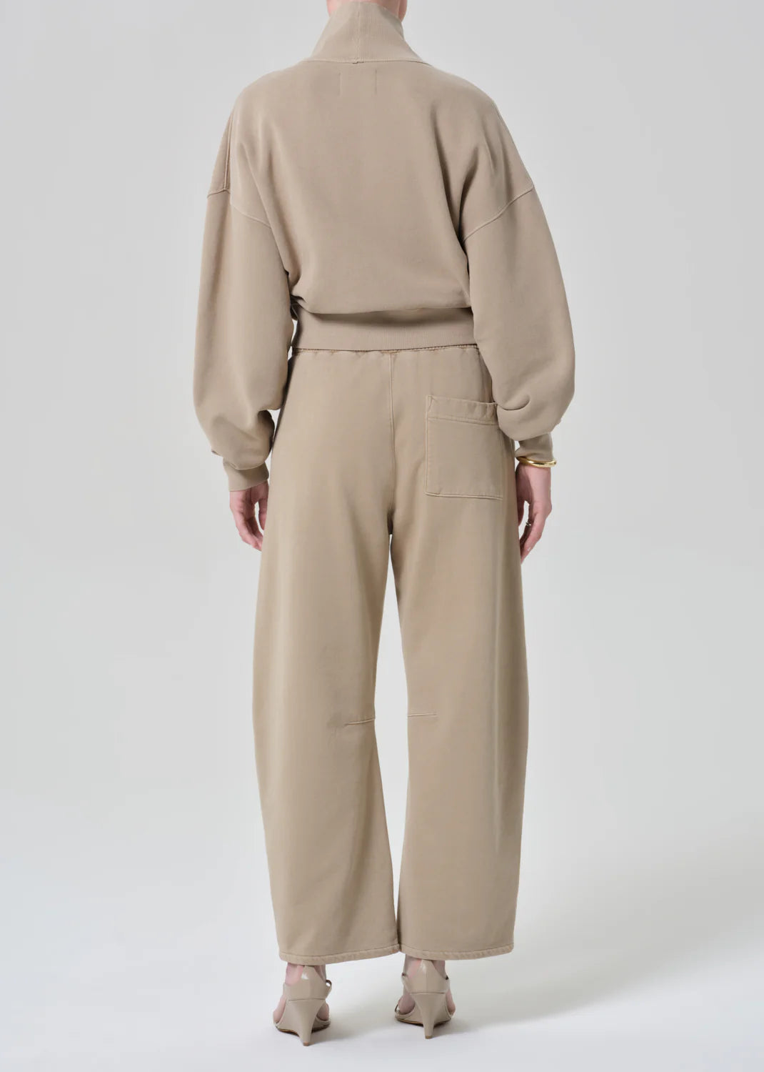 A person stands facing away, wearing Citizens Of Humanity/AGOLDE miro sweatpants with wide legs and cozy fit, paired with a beige jumpsuit and heeled shoes against a plain light background.