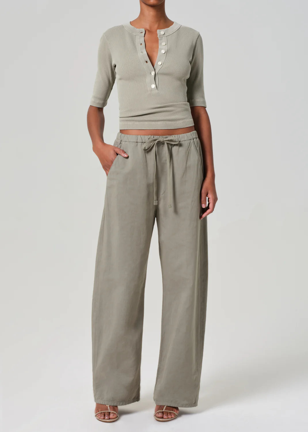 A person is wearing a Nali Short Sleeve Henley by Citizens Of Humanity/AGOLDE, paired with high-waisted, wide-leg drawstring pants and open-toed shoes made of organic cotton, standing against a plain background with one hand in their pocket.