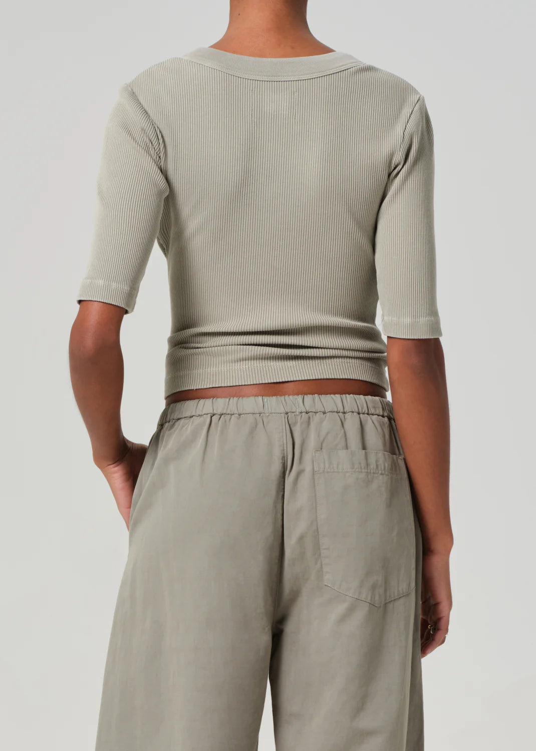 A person is seen from the rear wearing the Citizens Of Humanity/AGOLDE Nali Short Sleeve Henley in ribbed light gray with elbow-length sleeves, paired with high-waisted, loose-fitting pants of organic cotton against a plain white background that enhances the outfit's natural elegance.