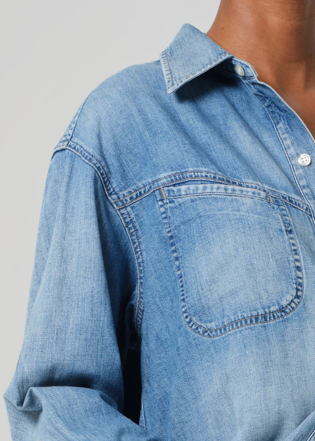 Close-up of a person wearing the Solene Shirt by Citizens Of Humanity/AGOLDE, featuring light blue Japanese denim with a pointed collar and visible front pocket. The fabric has a slightly worn texture, with the top button undone against a neutral gray background.