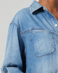 Close-up of a person wearing the Solene Shirt by Citizens Of Humanity/AGOLDE, featuring light blue Japanese denim with a pointed collar and visible front pocket. The fabric has a slightly worn texture, with the top button undone against a neutral gray background.