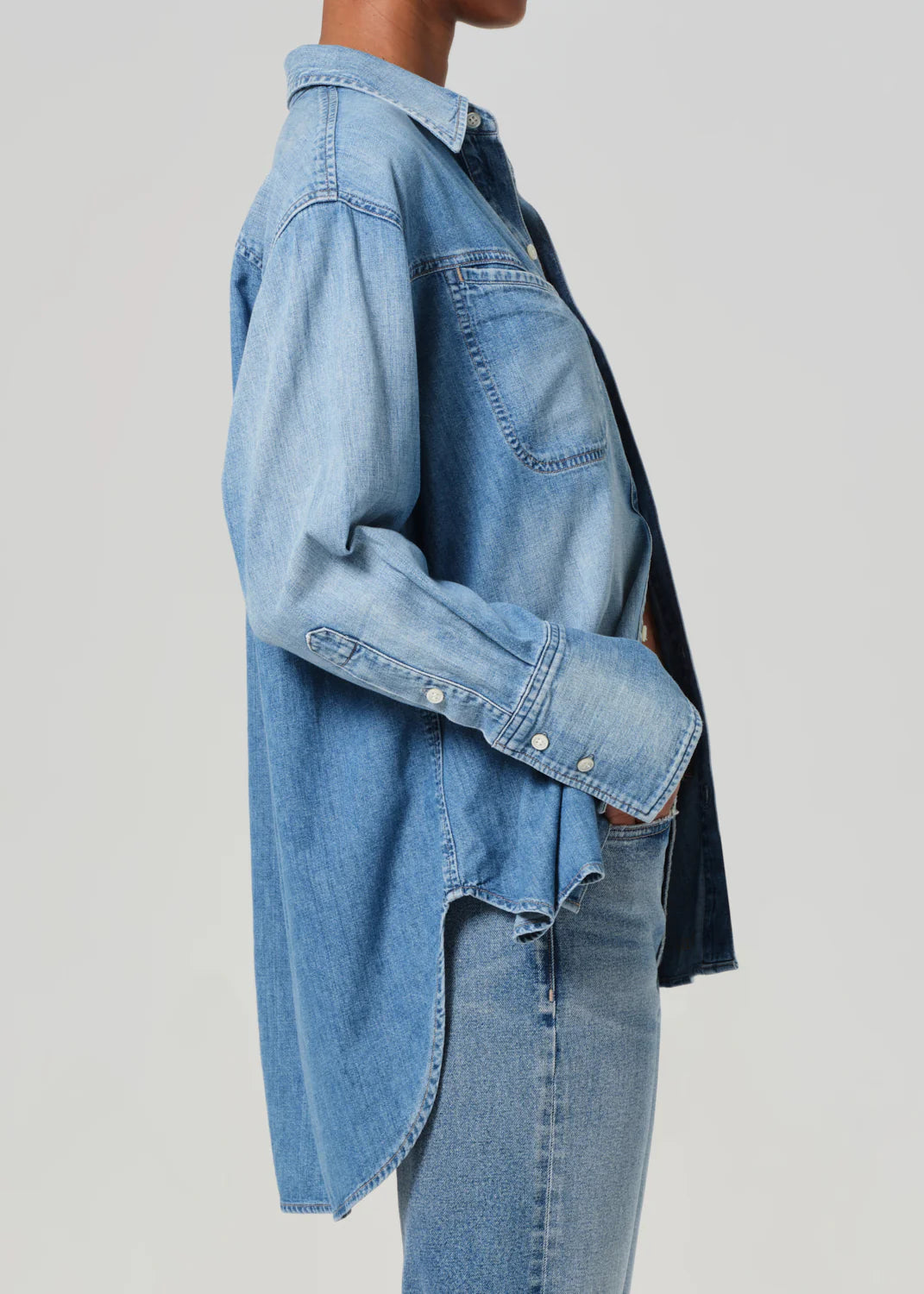 A person wearing the Solene Shirt, an indigo long sleeve denim piece from Citizens Of Humanity/AGOLDE, and jeans stands sideways with their hand in their pocket. The high-quality Japanese denim shirt features buttons and front pockets against a plain light gray background.