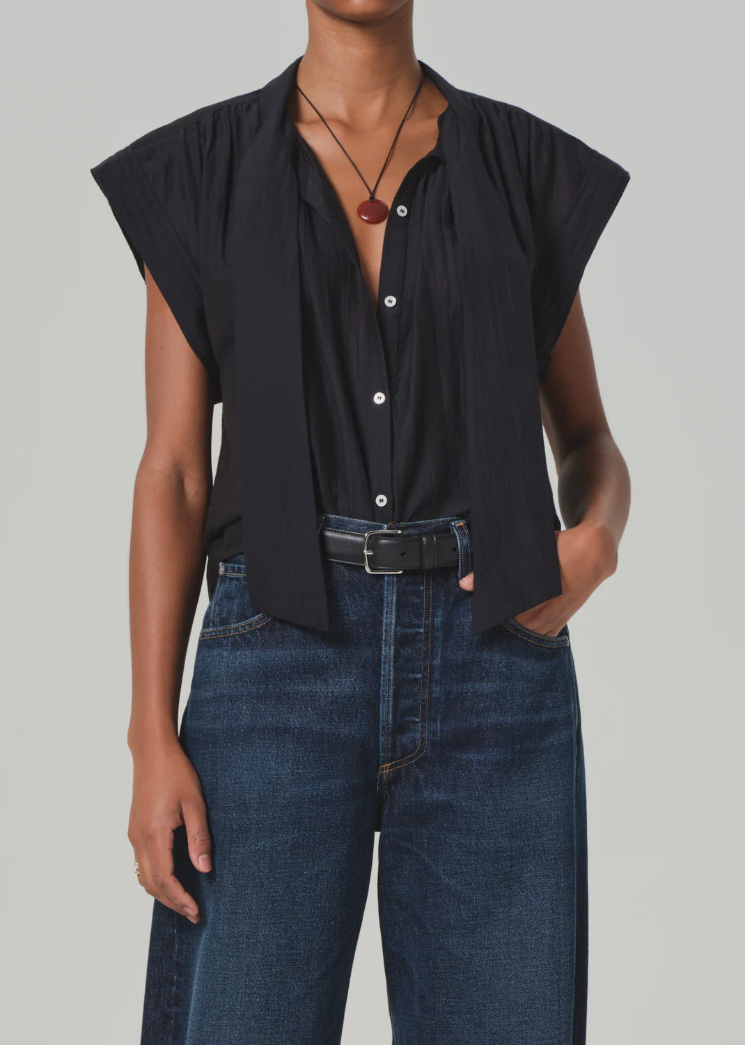 A person wearing the Penny Blouse with neck tie by Citizens Of Humanity, paired with high-waisted dark jeans from AGOLDE and a black belt. They accessorize with a thin necklace featuring a circular pendant, hand in pocket against a light gray background.
