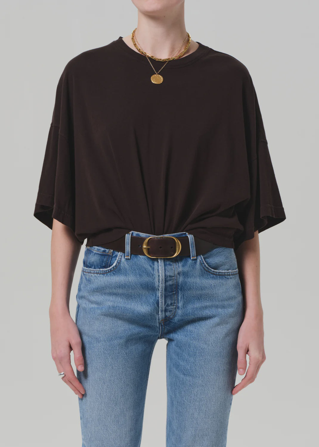 A person wears the Citizens Of Humanity/AGOLDE Cape Tee in a loose, boxy fit tucked into high-waisted blue jeans, accessorized with a black belt with gold buckle and layered gold necklaces. The background is plain and light-colored.