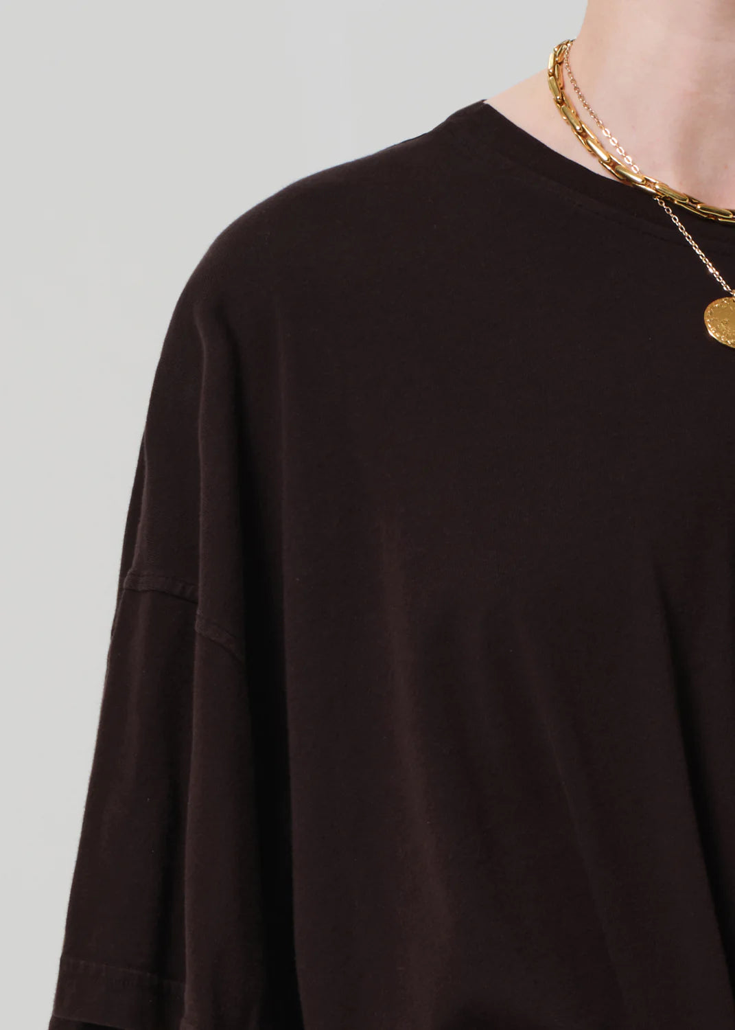 A close-up shows a person wearing the Cape Tee by Citizens Of Humanity/AGOLDE—a boxy fit black T-shirt made from regenerative cotton—styled with layered gold necklaces, one featuring a round pendant, against a light neutral background.