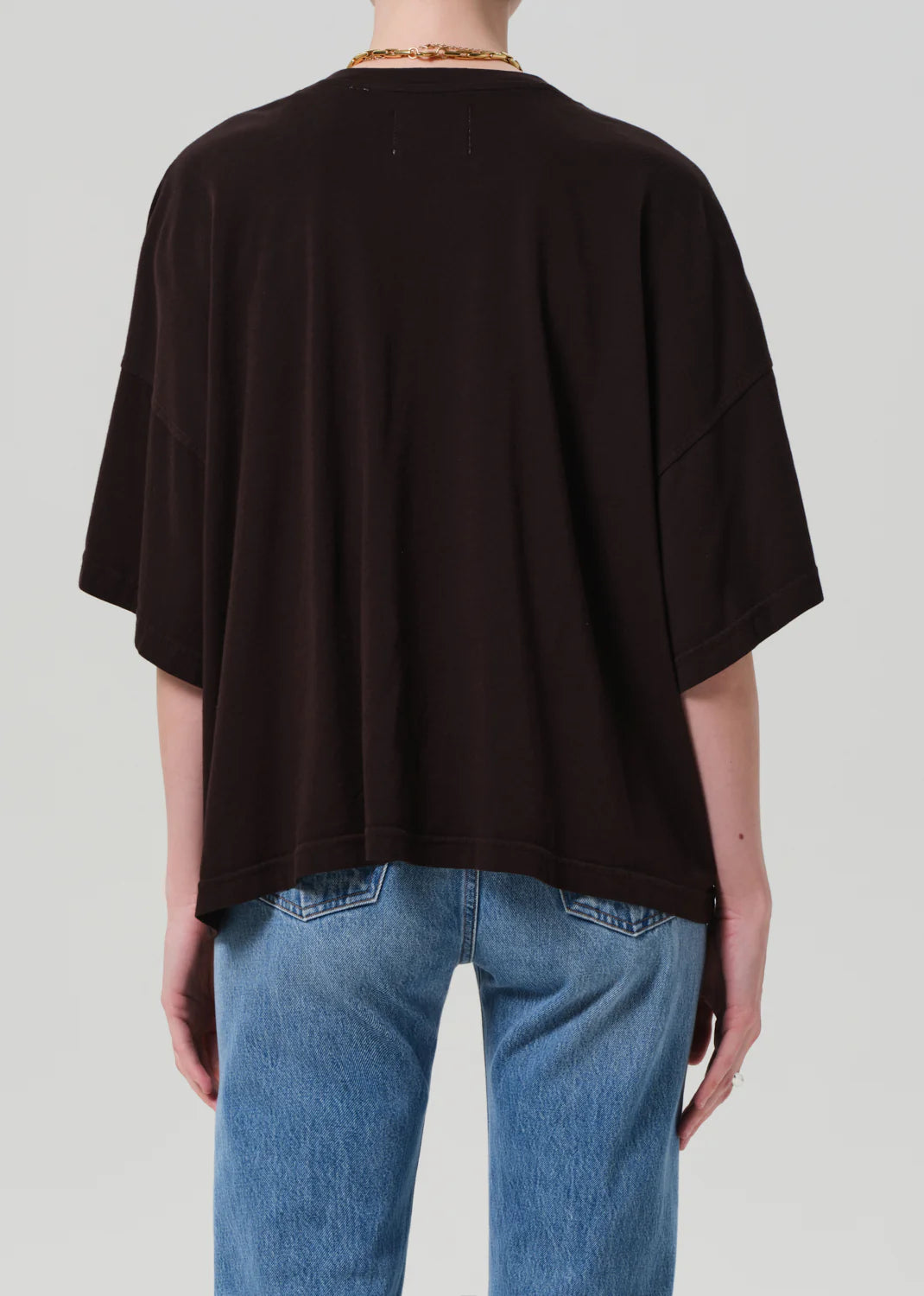 A person is shown from the back against a plain light background wearing the Citizens Of Humanity/AGOLDE Cape Tee, a loose-fitting black T-shirt made from regenerative cotton with wide sleeves, a boxy fit, and effortless style, paired with blue jeans.