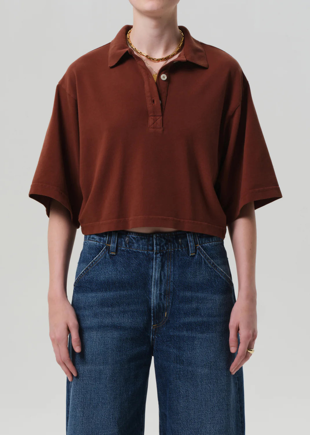 A person wears the Lowen Polo Shirt by Citizens Of Humanity/AGOLDE in brown supima cotton, styled oversized with high-waisted dark blue jeans. Gold necklace and rings complete the look against a plain light background.