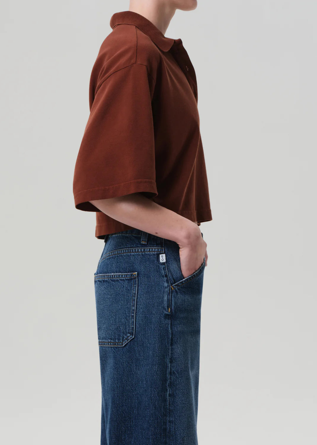 A person stands in profile with one hand in their pocket, wearing the oversized brown Lowen Polo Shirt by Citizens Of Humanity/AGOLDE and blue high-waisted jeans against a plain light background.