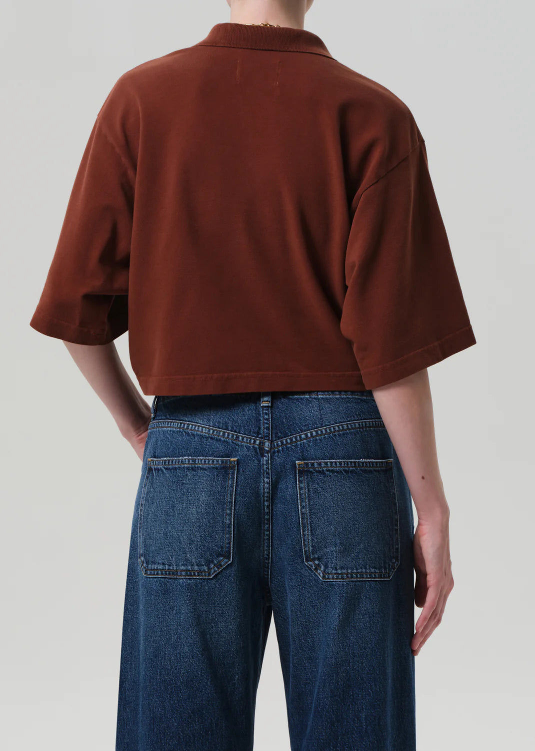 A person faces away, wearing a loose-fitting brown Lowen Polo Shirt by Citizens Of Humanity/AGOLDE with elbow-length sleeves and high-waisted blue jeans with visible back pockets, set against a neutral light gray background.