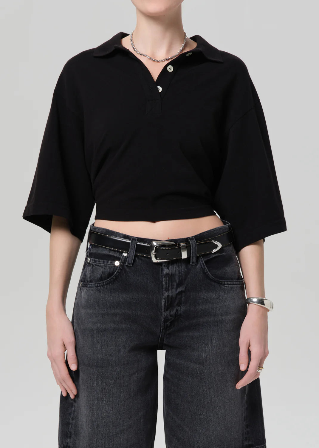 Someone is wearing the lowen polo shirt by Citizens Of Humanity/AGOLDE, paired with black high-waisted jeans, a black belt, and silver jewelry against a plain light background.